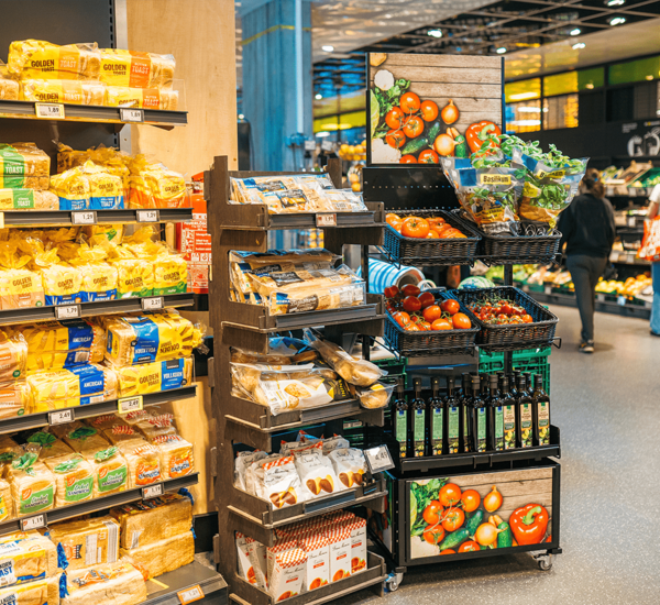 Différentes sortes de légumes, comme par exemple des tomates et des herbes aromatiques, présentées dans des paniers avec un deuxième point de vente. Deux clientes sont visibles à l'arrière-plan.