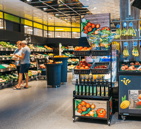 Deuxième point de vente avec trois tablettes et un présentoir avec une finition bois « impression légumes », différents paniers avec des légumes et des herbes aromatiques, de l'huile d'olive est présentée sur une tablette, des clients en arrière-plan.