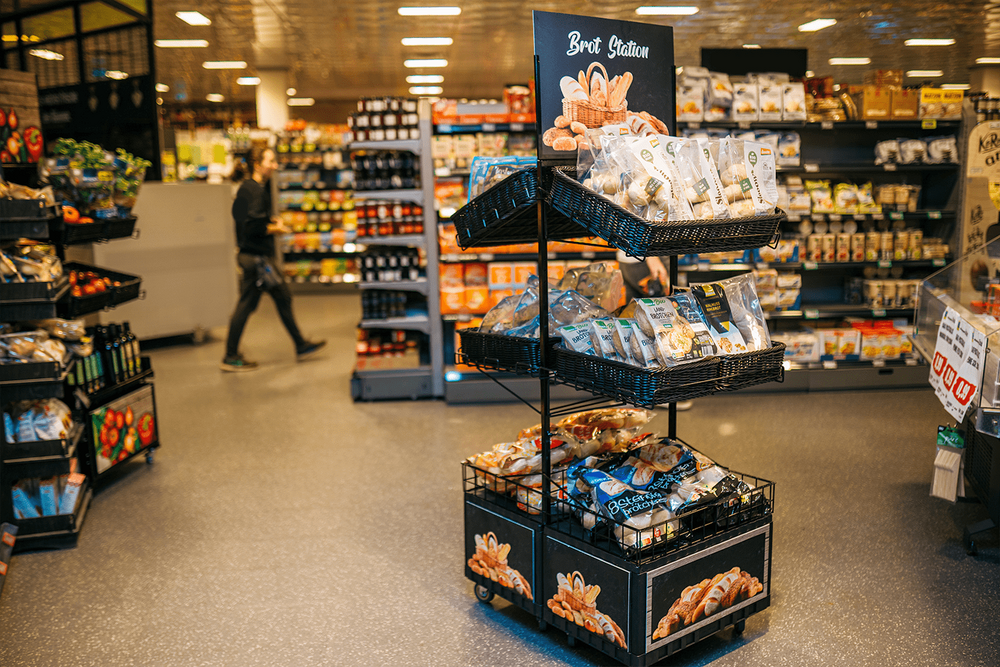 Zweiter Point of Sale Doppelseitig mit 6 Tablaren und Topper (Aufschrift Brot Station, Abbildung Brotkorb mit divers. Gebäck), auf den 6 Tablaren sind unterschiedliche Aufbackbrote präsentiert. Im Hintergrund Kunde ersichtlich.