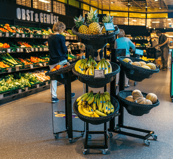 Deuxième point de vente avec six paniers, trois paniers (prise de vue frontale) contiennent différents fruits comme des bananes et des ananas. Clients à l'arrière-plan. Une cliente sur le côté.