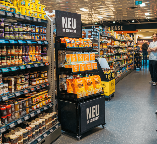 Deuxième point de vente avec trois tablettes et un présentoir portant l'inscription « NOUVEAU DANS L'ASSORTIMENT ». Différentes sortes de biscottes et de pains croustillants sont présentées. À côté, une étagère avec diverses pâtes à tartiner.